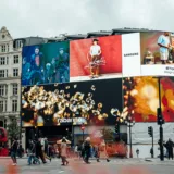 Scena diurna di Piccadilly Circus a Londra, con grandi schermi DOOH che mostrano pubblicità di vari marchi come Samsung, Rabanne e Louboutin. Persone e autobus rossi attraversano la strada affollata.