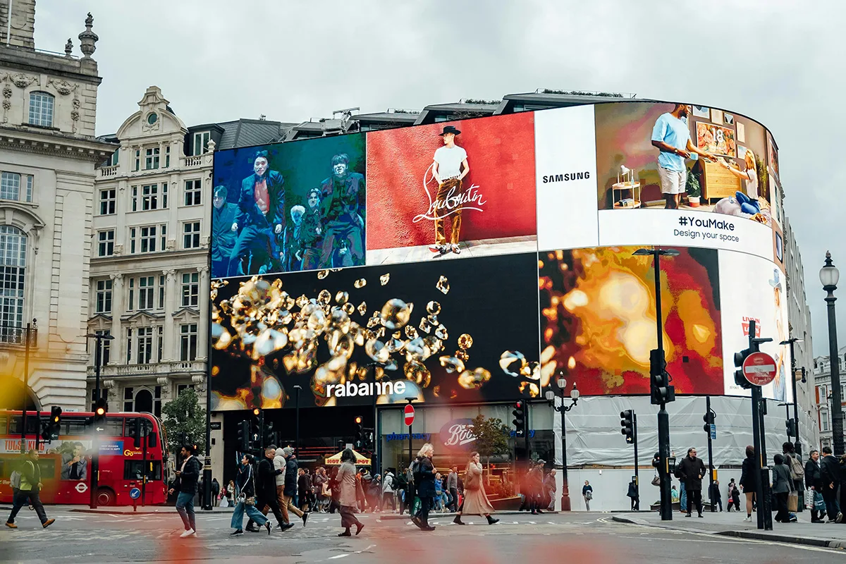 Scena diurna di Piccadilly Circus a Londra, con grandi schermi DOOH che mostrano pubblicità di vari marchi come Samsung, Rabanne e Louboutin. Persone e autobus rossi attraversano la strada affollata.