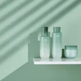 Presentazione minimalista di prodotti Hanyul per la cura della pelle in confezioni verde chiaro su uno scaffale bianco, con ombre di luce solare che creano un motivo a strisce sullo sfondo verde