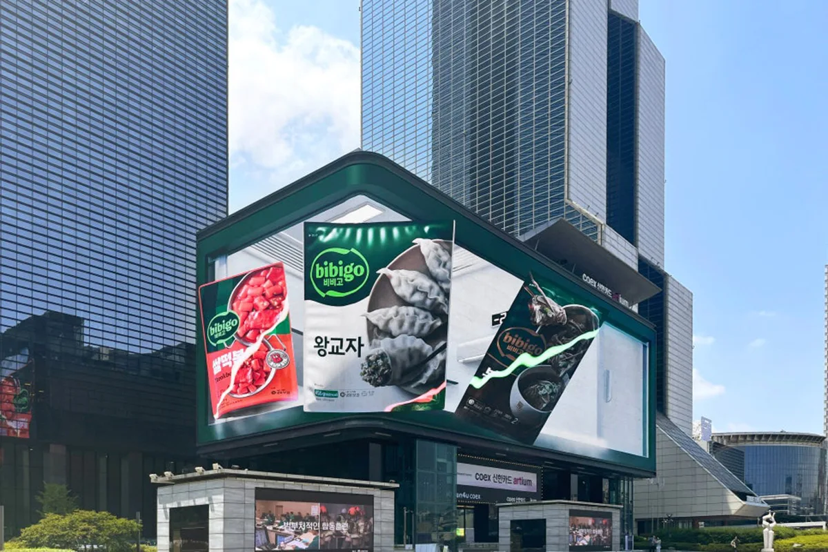 Grande schermo DOOH a Seoul, Corea, che mostra una pubblicità di Bibigo con prodotti come ravioli e zuppe, sullo sfondo di alti edifici moderni in una giornata di sole.