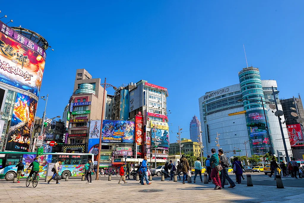 Affollata via di Taipei, Taiwan, con schermi DOOH che mostrano pubblicità colorate su edifici e un autobus che passa. La scena è vivace con pedoni e ciclisti sotto un cielo azzurro.